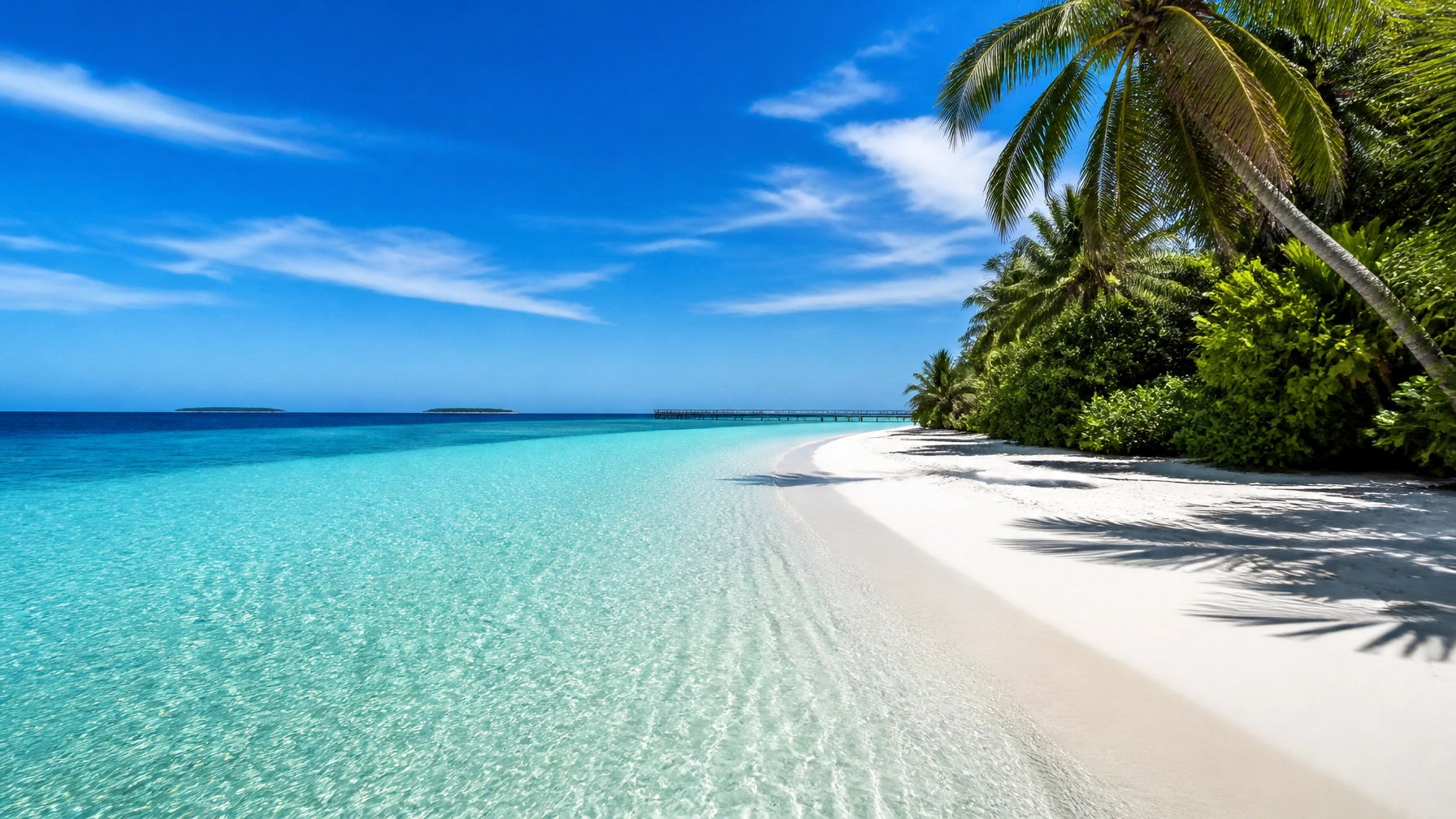 A stunning, wide-angle hero shot of a remote island in the Southern Maldives. The water is crystal clear turquoise, fading into deep sapphire. A pristine, white sand beach stretches along the shore with no people in sight.
